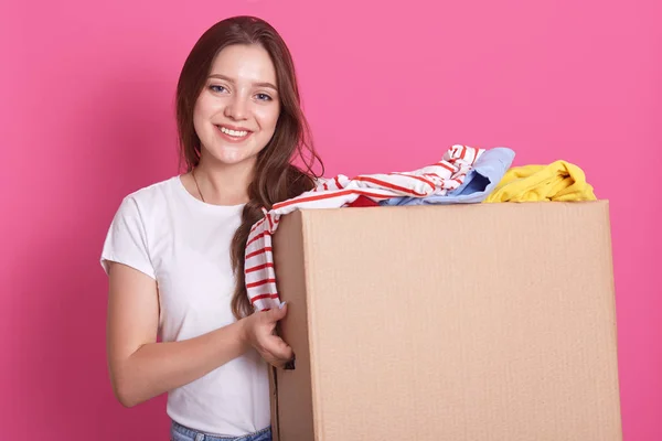 Horizontal shot of female stands smiling near both with reusable clothing for poor peoplw isolated over pink background in studio, attractive woman making charity, posing with happy expression.