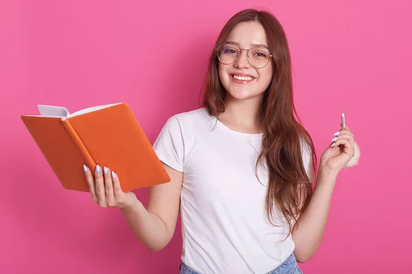 Plano horizontal de joven atractiva mujer caucásica en ropa blanca casual y gafas, sosteniendo portátil y penil en las manos, modelo posando aislado sobre la pared del estudio de color rosa. Concepto de estudiantes . —  Fotos de Stock