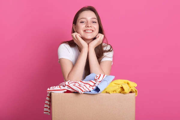 Cheerful helpful volunteer standing isolated over pink background in studio, holding box with clothes for secondary use, keeps hands under chin, looks at camera with pleased facial expression. — Stock Photo, Image