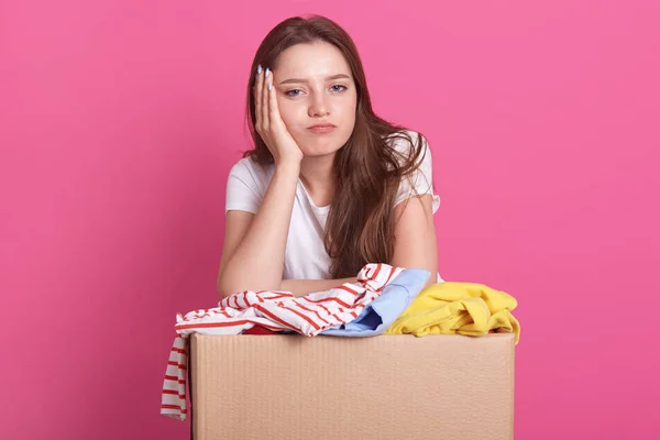 Close up portrait of happy young woman with clothes donation standing over rose background, packing reusable clothes for poor people, attractive female keeps hand under chin, looking bored at camera. — Stock Photo, Image