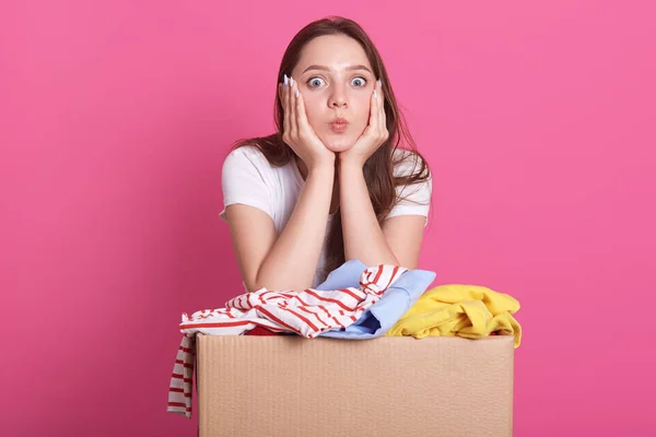 Studio shot of cheerful volunteer standing isolated over pink background in studio, near box with donated clothes, young attractive woman makes air kiss, looks directly at camera. Charity concept. — Stock Photo, Image