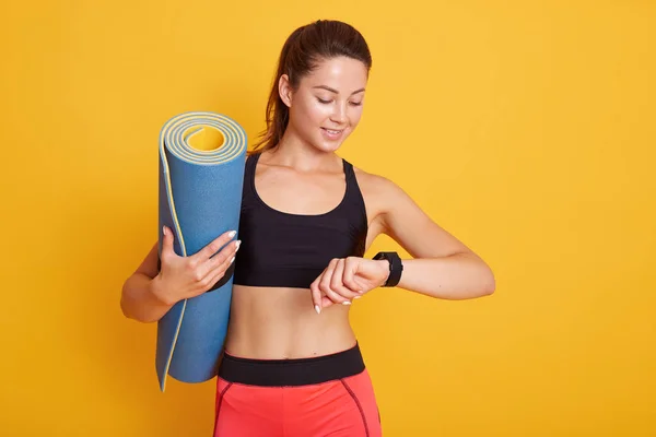 Horozontal shot of fitness woman after workout session checks results on smartwatch in fitness app, female with pefect body isolated over yellow background. Estilo de vida saludable y concepto deportivo . — Foto de Stock