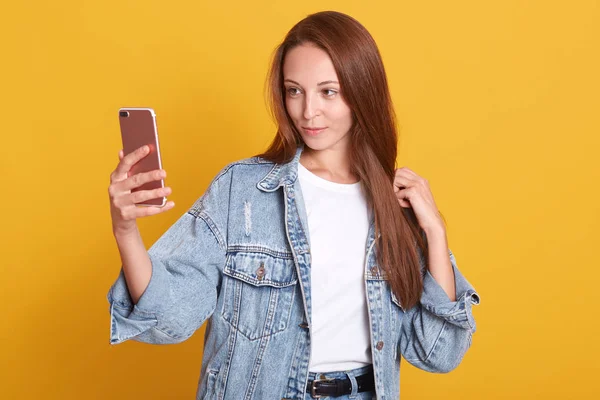 Gambar gadis berkaos putih dan jaket demin yang sedang selfie terisolasi dengan latar belakang kuning memegang smartphone dengan tangan, melihat pada perangkat layarnya, wanita dengan rambut panjang dan lurus . — Stok Foto