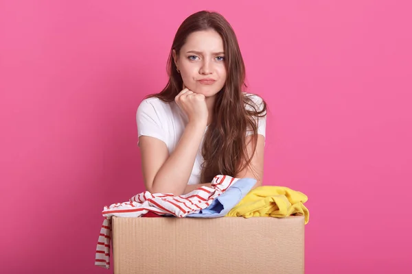 Close up portrait of beautiful woman with pensive facial expression, posing near box with old clothes, decides whom to give things for secondary use, standing over pink background. Donation concept. — Stock Photo, Image