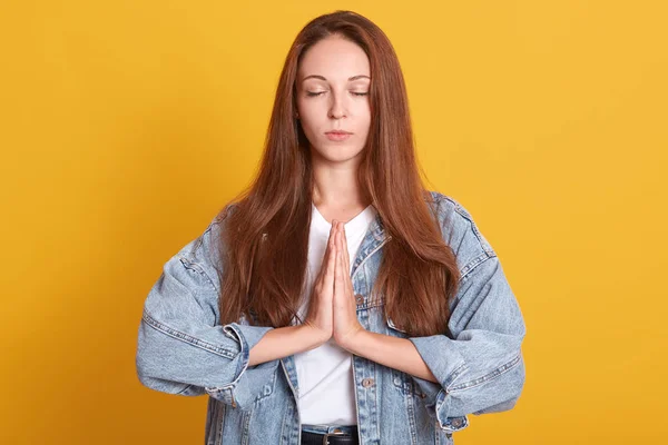 Foto interior de atractiva mujer europea vestida con chaqueta de mezclilla cogida de la mano en oración, de pie con los ojos cerrados sobre la pared amarilla. Se ve calmado, con el pelo largo y claro. Copiar espacio con advertismant — Foto de Stock