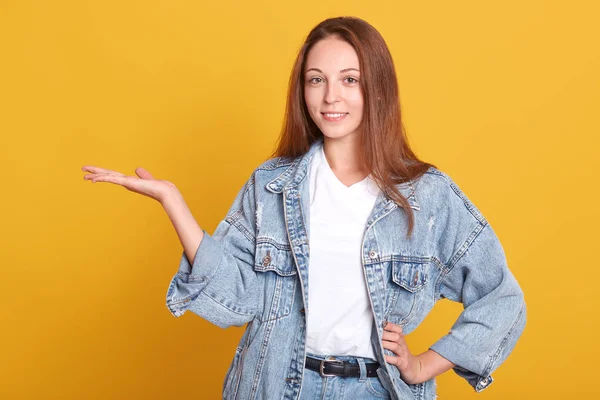 Mulher adorável positivo com hiar satraight escuro, mantém a palma levantada, demonstra spmething, estar em alto espírito, posando sorrindo isolado sobre fundo amarelo. Copiar espaço para promoção ou adicionar . — Fotografia de Stock