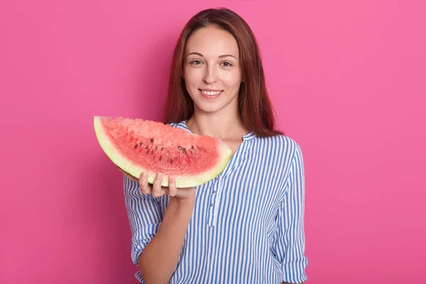 Foto interior de jovem com grande pedaço de melancia na mão, mulher adorável vestindo camisa listrada, olhando sorrindo para a câmera, modelo posando isolado sobre fundo de estúdio rosa. Conceito de pessoas . — Fotografia de Stock