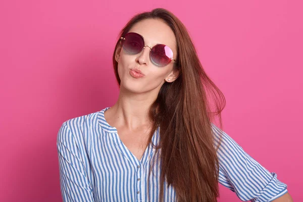 Indoor shot de mujer encantadora con gafas de sol con estilo y camisa a rayas, mantiene los labios redondeados, haciendo gesto beso, modelo posando aislado sobre fondo de estudio rosa. Gente emociones concepto . —  Fotos de Stock