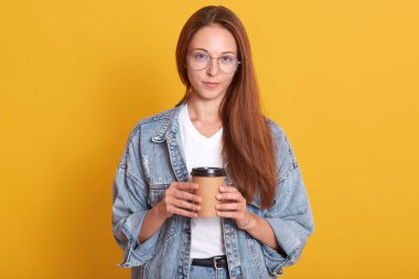 Neşeli bayan zevkle kameraya doğrudan bakarak stüdyo portresi, sıcak kahve tutan genç kadın, şık kot ceket giyen, sarı arka plan üzerinde izole poz. Yaşam tarzı kavramı.