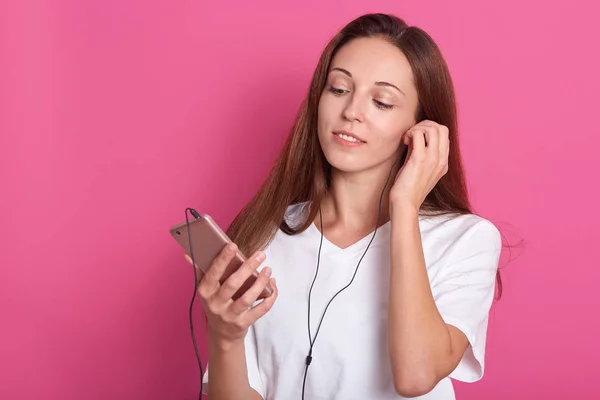 Wanita ikat menempatkan earphone di telinga. Potret gadis pemimpi yang santai dan menarik dengan rambut yang panjang, melihat layar telepon pintar, mendengarkan musik, mengenakan pakaian santai . — Stok Foto