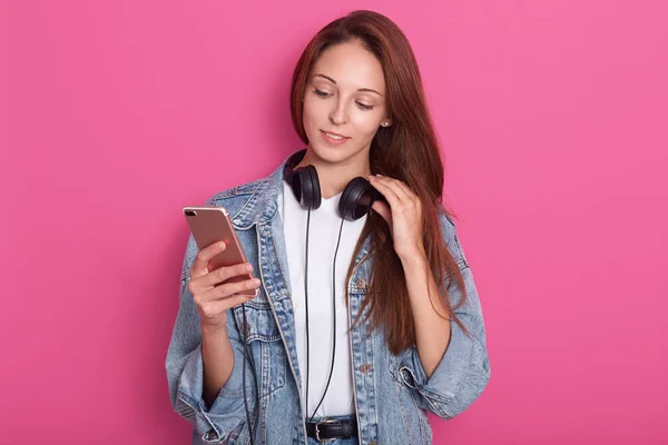 Young attractive girl standing isolated over pink background, holding her smartphone in hand, having headphones around neck and touches it with her fingers, looks at her device, wearing stylish outfit — Stock Photo, Image