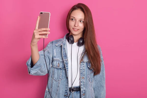 Vakker jente som tar selfie mot rosa studiovegg. Smilende kvinne i stilig dongerijakke og hvit skjorte, som holder smarttelefon og lytter til musikk via øretelefoner, ser på apparatet sitt . – stockfoto
