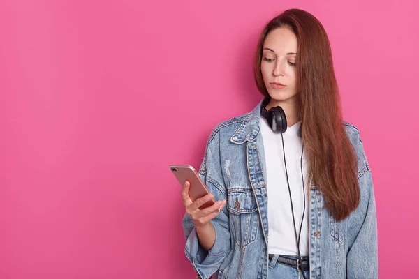 Junge schöne Frau mit Smartphone, während sie isoliert vor rosa Hintergrund posiert, Jeansjacke trägt, Kopfhörer um den Hals hat, das Gerät betrachtet. Kopierfläche für Werbung oder Promotion. — Stockfoto
