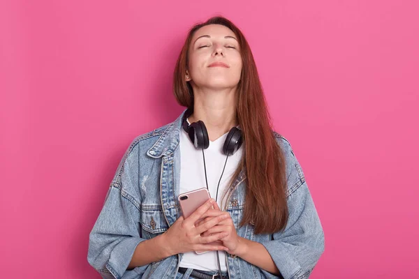 Porträt eines jungen Mädchens in stylischer Jeansjacke, das Handy in den Händen hält, mit geschlossenen Augen den Kopf hochhält, dankbar ist, froh, die Nachricht vom Freund zu hören, isoliert über rosa Hintergrund. — Stockfoto
