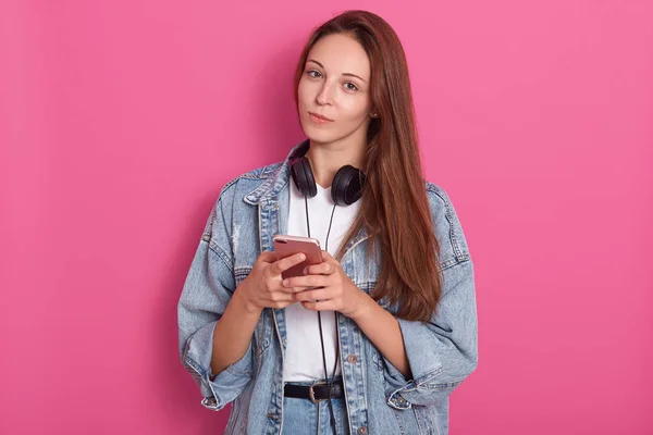 Charming woman with dar hair holding smartphone in both hands and looking at the camera over rose background, dressed stylish outfit, has head phones around neck, like to listen music, looks serious. — Stock Photo, Image