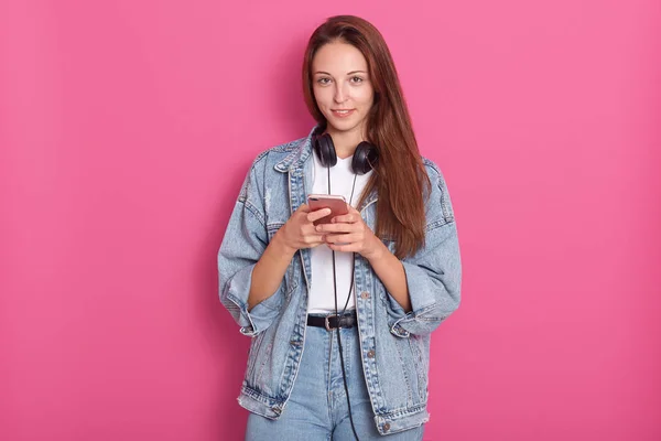 Retrato de una joven hermosa y feliz usando su teléfono, mirando directamente a la cámara y sonriendo, usando chaqueta de mezclilla, camisa blanca y jeans, con cabello largo y liso, aislado sobre fondo rosa . — Foto de Stock