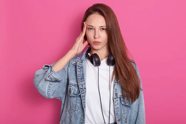 Indoor shot van jonge vrouw in denim casual kleding, hoofdtelefoons rond nek, heeft hoofdpijn van luide muziek, ziet er moe uit, houdt vinger op de tempel, geïsoleerd op roze oranje achtergrond in Studio. — Stockfoto