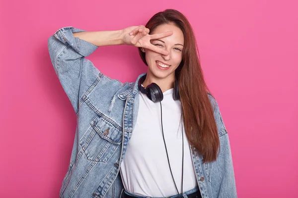 Front view of beautiful happy young woman looking directly at camera, holding peace sign near eye and standing isolated over rosy background, wearing denim jacket, holding headphones around neck. — Stock Photo, Image