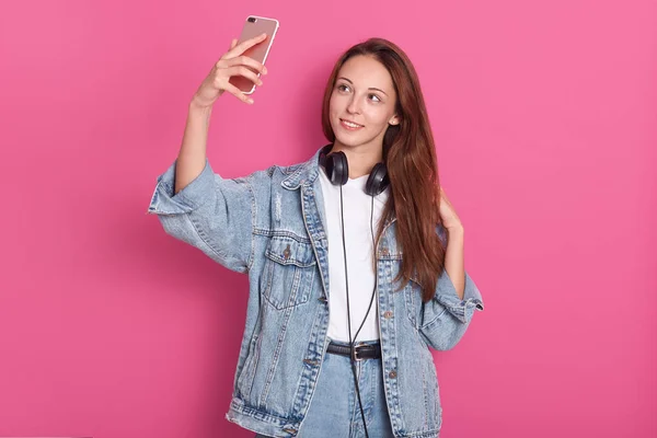 Porträt einer hübschen jungen Frau in Jeansmode, die ein Selfie mit dem Handy macht, Kopfhörer um den Hals trägt, lange Haare hat und isoliert auf rosa Hintergrund im Studio posiert. — Stockfoto