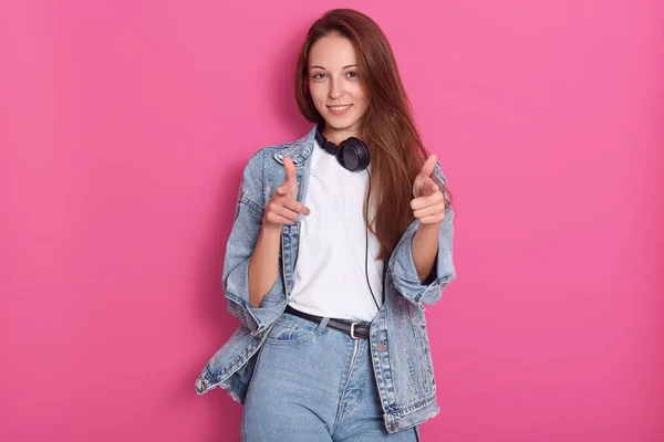 Positiv munter ung kaukasisk kvinde med langt hår, iført hvid afslappet t-shirt, jeans og denim jakke, holder hovedtelefoner rundt om halsen, peger på kameraet med pegefingrene og smiler . - Stock-foto