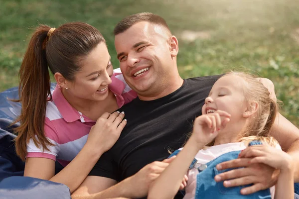 Keluarga muda yang bahagia menghabiskan waktu luang di taman untuk piknik, ibu dan ayah tertawa karena lelucon donatur, mereka berpakaian santai, memiliki ekspresi wajah yang bahagia. Konsep keluarga, hubungan dan kebahagiaan . — Stok Foto