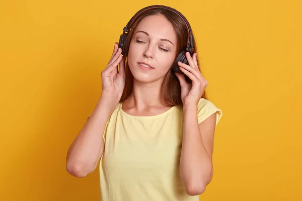 Lukk opp portrettet av en vakker, kaukasisk kvinne som hører på musikk via hodetelefoner, slapper av mens hun har tid, poserer med lukkede øyne, kler seg uformelt, isolert over gul studiobakgrunn – stockfoto