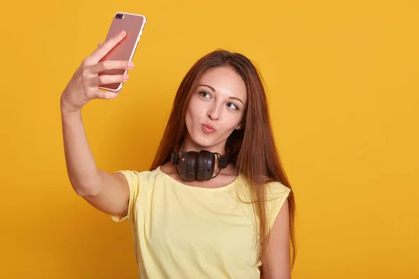 Nært portrett av en vakker, fantastisk dame som lager selfie via telefonen, tar bilde til kjæresten, går i vanlig t-skjorte, har langt hår, poserer isolert over lys gul bakgrunn . – stockfoto