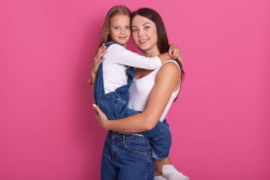 Küçük kızı kucaklayan ve pembe arka plan üzerinde izole gülümseyen genç sevimli anne Fotoğrafı, rahat kıyafet giyen, doğrudan kameraya bakarak, mutluluk ifade. Aile kavramı.