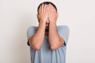 Genç adam rahat gri t shirt giyen görüntü üzücü ifade iki eliyle yüz kapsayan izole beyaz stüdyo arka plan üzerinde duran, sorunlar yaşıyorsanız. Depresyon sonu duygular kavramı.