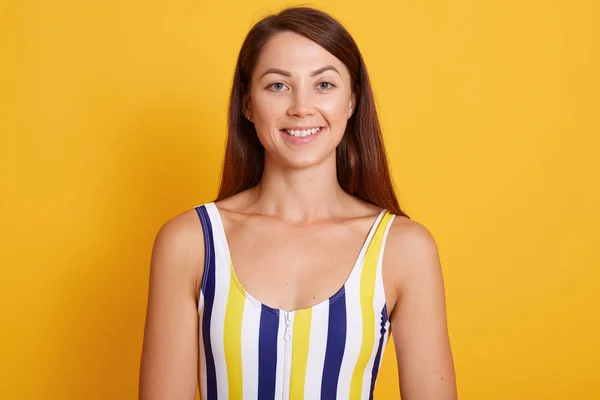 Hermosa mujer de pelo oscuro vestido traje de baño a rayas está sonriendo y mirando directamente a la cámara. Estudio plano de encantadora hembra posando aislada sobre fondo amarillo. Concepto de personas . — Foto de Stock