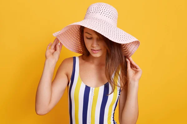 Femme caucasienne pensive portant un maillot de bain debout avec les mains sur le chapeau et regardant vers le bas, pleine de pensées, portrait demi-longueur d'une jolie femme, posant isolée contre un mur de studio jaune . — Photo