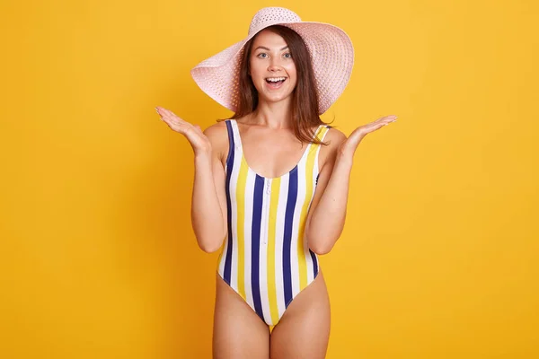 Primer plano retrato de mujer sorprendida en sombrero de verano y traje de baño a rayas, mirando con los ojos tapados y abre la boca de sorpresa, de pie sobre fondo amarillo. Gente emociones concepto . — Foto de Stock