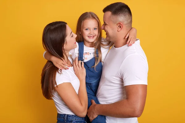 Happy young Caucasian family in photo studio. Father, mother and charming daughter. Parents hugging little adorable girl dressed denim overalls. Woman and man smiling and look at their child.