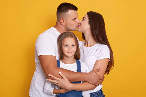 Una giovane famiglia felice. Marito baciare moglie e la loro graziosa figlia indossando tuta in denim guardando direttamente alla fotocamera, i genitori vestiti casual t shirt bianche. Studio ritratto su sfondo yello . — Foto Stock