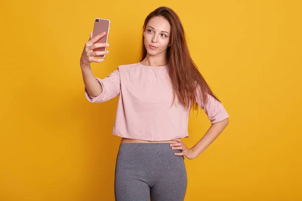 Студийный снимок красивой темноволосой женщины с прямыми волосами в серых брюках и розовой рубашке, делающей селфи с мобильным телефоном, держит свое устройство в руке, позируя изолированным на желтом фоне . — стоковое фото