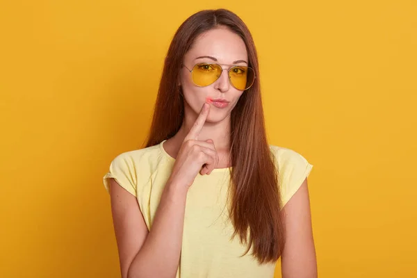Close up portrait of beautiful dark haired woman points to her lips with fore finger, has virus, being infected with col sore herpes, needs treatment, dressed casually, isolated over yellow background