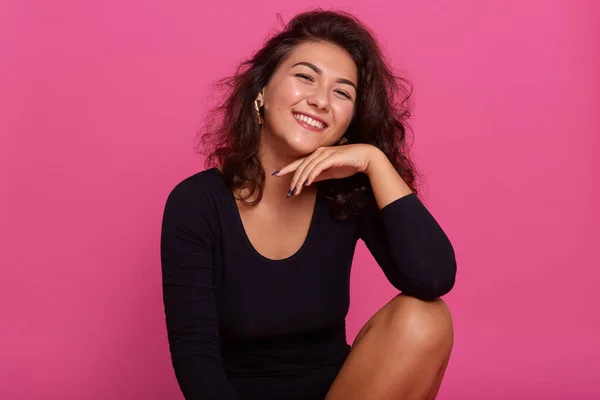 Retrato de cerca de la atractiva joven mujer que mantiene la mano bajo la barbilla, sonriendo mientras se sienta sobre el fondo rosa, vistiendo camisa negra, estar de buen humor, tener el pelo ondulado oscuro y pendientes con estilo . —  Fotos de Stock