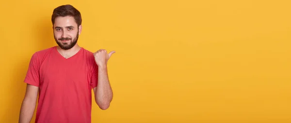 Obraz optimistického hezkého muže, který ukazoval stranou palcem a dívá se na kameru, oblečený v červeném, neformální košili, kopírovací prostor pro propagační text nebo inzerci, izolované přes žluté pozadí studia. — Stock fotografie