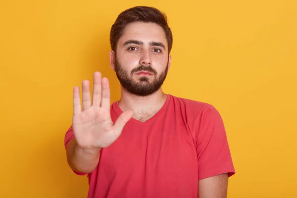 Kırmızı rahat tshirt giyen sakin sakallı genç adam portresi uyarı jest el ile ayakta ve ciddi bir yüz ile kameraya bakarak, yakışıklı erkek sarı arka plan üzerinde izole poz. — Stok fotoğraf