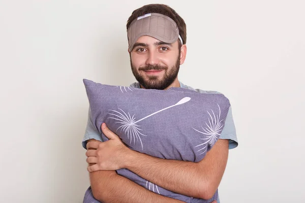 Indoor tiro de homem bonito jovem fica sorrindo e abraçando travesseiro, macho posando no quarto com venda na testa, olhando diretamente para a câmera, tem aparência agradável, pronto para dormir . — Fotografia de Stock