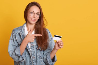 Gülümseyen kadın altın kredi kartı tutan ve onun işaret parmağı ile işaret, advertismant veya tanıtım metin için kopya alanı, kadın şık kot ceket ve gözlük giyen, sarı duvarda izole.