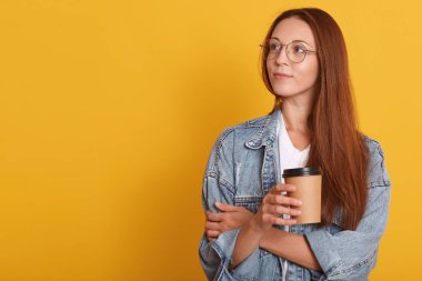 Koyu saçlı düşünceli kadın yakın portresi, kenara bakarak, bir şey hakkında düşünüyor, beyaz t shirt giyiyor, kot ceket abd gözlük, kahve tek kullanımlık fincan tutan, sarı duvar üzerinde.