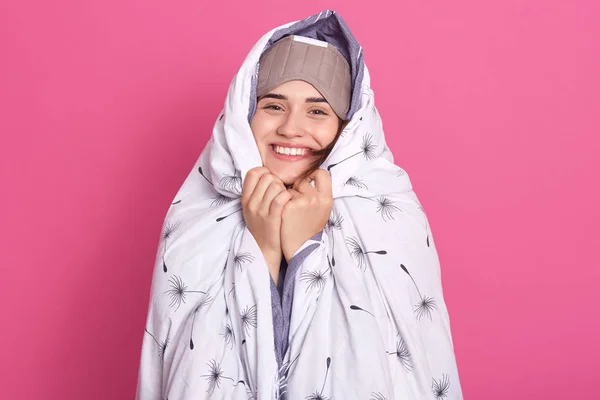 Smiling woman wakes up in morning, posing with blanket and blindfold eyes mask for sleep, having happy facial expression, has weekend, posing isolated over pink studio background. Relaxing concept.