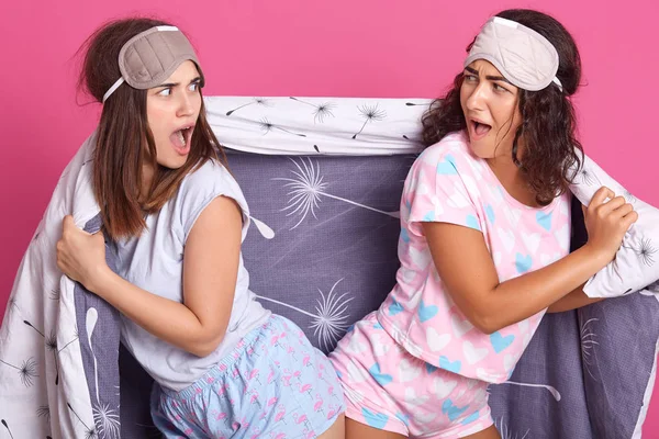 Horizontal shot of unhappy girls posing isolated over pink background, females standing back to back, crying something, having quarrel, ladies wearing pajamas and blindfolds. Friendship concept.