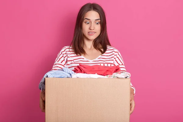 Horizontal shot of young volunteer stands with carton box full of donated clothes, adorable woman looking at clothes with scared facial expression, bits her low lip, lady wearing striped casual shirt. — Stock Photo, Image