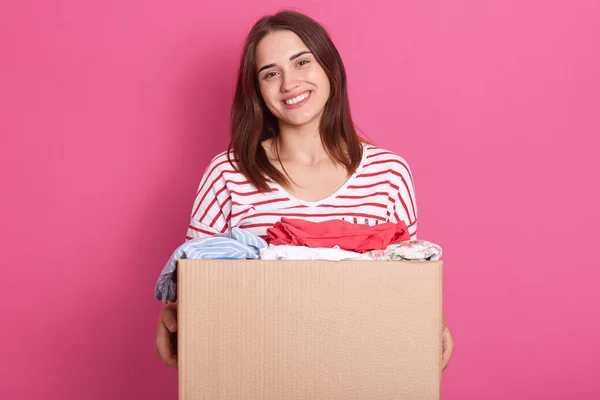 Horizontal shot of happy female posing isolated over pink background, holding carton box with rausable clothes, clothing for poor people, charming woman making charity, dressed striped shirt.