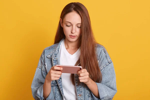 Close up portrait of young beautiful woman using smart phone, writting message to her friend, wearing denim jacket and white casual t shirt, looking at device, looks concentrated. People concept. — Stock Photo, Image