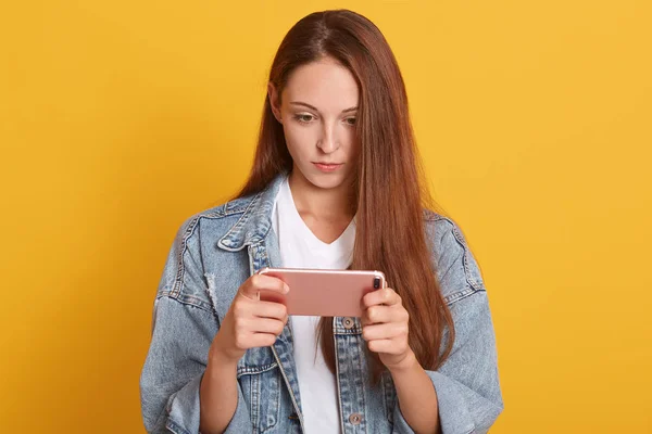 Image of attractive concentrated woman holding cell phone in hands, female wering denim jacket and white t shirt, looks at device with serious facial expression, posing isolated over yellow background — Stock Photo, Image