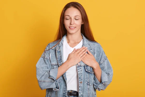 Närbild porträtt av vacker kvinna bär snygg jeansjacka och vit t-shirt, stående över isolerad gul bakgrund, stående med händerna på bröstet med slutna ögon och tacksam gest på ansiktet. — Stockfoto