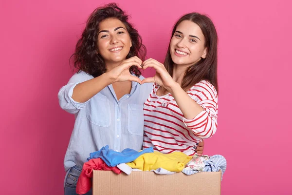 Pleasant young women wearing casual clothes posing isolated over pink background in studio. Females showing shape heart with hands, expressing love, looking smiling at camera. People concept. — Stock Photo, Image
